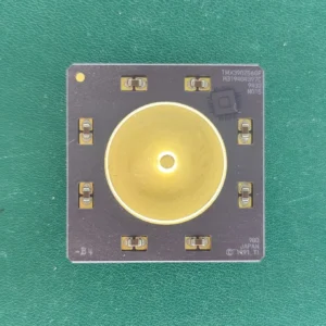 Top view of TI TMX390Z566F ceramic hybrid module with a gold-plated central disc on a green background.